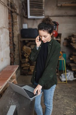 Esmer marangoz akıllı telefonla konuşuyor ve atölyede dizüstü bilgisayar kullanıyor. 