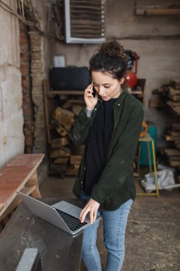Brunette woodworker talking on smartphone and using laptop in workshop  clipart