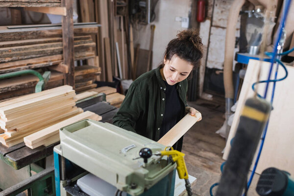 Brunette carpenter holding wooden board near thickness planer and planks in workshop 