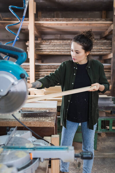 Woodworker holding plank near blurred miter saw on foreground in workshop 