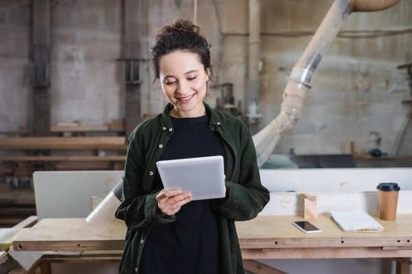 Smiling furniture designer using digital tablet near smartphone and safety visor in workshop 