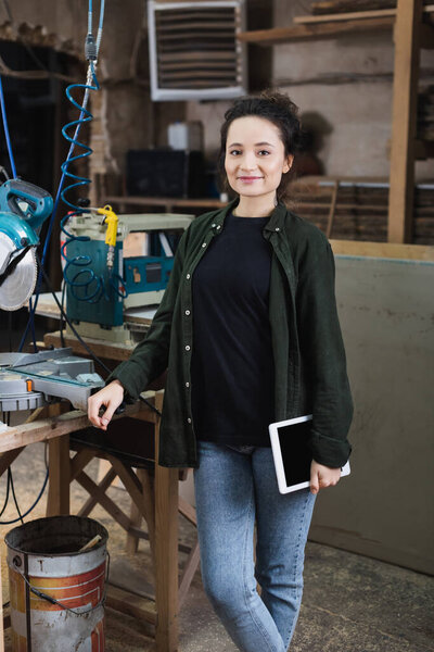 Positive carpenter holding digital tablet near miter saw in workshop 