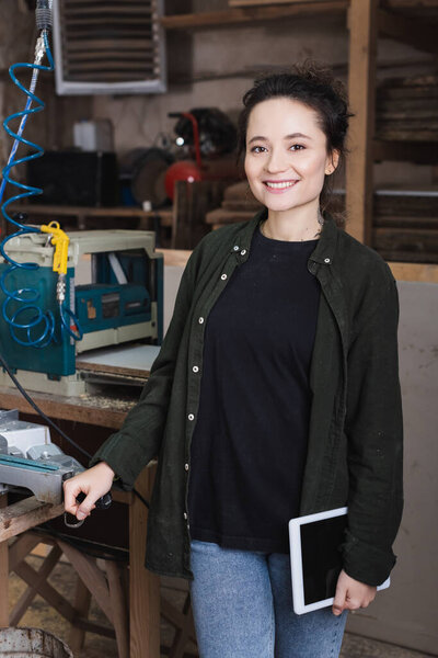 Cheerful furniture designer holding digital tablet near equipment in workshop 
