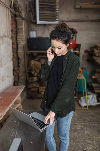 Brunette woodworker talking on smartphone and using laptop in workshop 