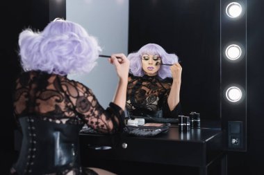 man in purple wig applying makeup near mirror in dressing room clipart