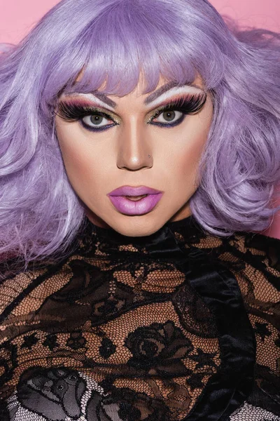 man in makeup, purple wig and black lace clothing looking at camera isolated on pink