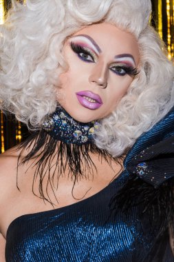 Muhteşem makyajlı travesti parlak arka planda kameraya bakıyor.