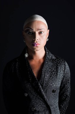 Piercingli ve yarı makyajlı bir adam siyah kameraya bakıyor.