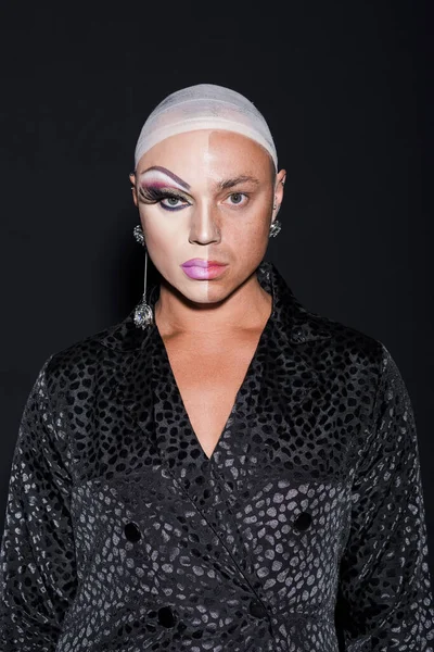 eccentric man with piercing and makeup on half face looking at camera isolated on black