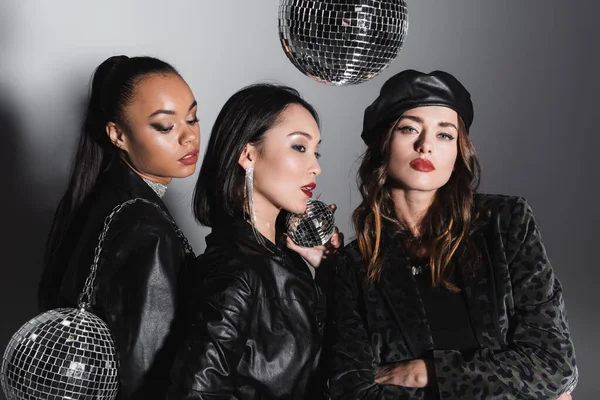 young woman in leather beret looking at camera near interracial models holding shiny disco balls on grey