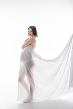 Bez giymiş hamile bir kadın beyaz arka planda kameraya bakıyor. 