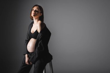 Koyu gri arka planda sandalyenin yanında poz veren ceketli moda hamile kadın.