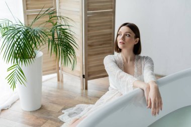 Young woman in robe looking away near bathtub and plant in bathroom  clipart