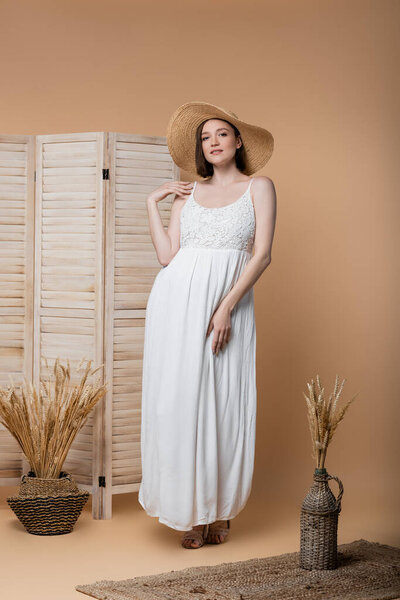 Стильная беременная женщина в соломенной шляпе позирует на бежевом 