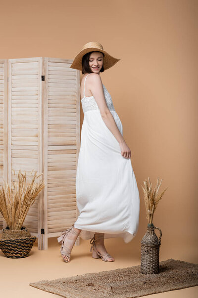 Счастливая беременная женщина трогает платье рядом с растениями и складной экран на бежевом 
