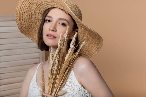 Young woman in straw hat holding spikelets near folding screen isolated on beige 