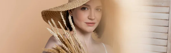 Woman in sun hat looking at camera near spikelets and folding screen isolated on beige, banner