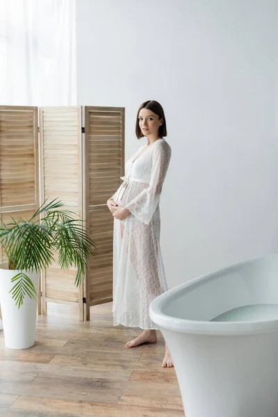 Pregnant woman in robe standing near plants and bathtub at home