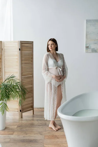 年轻孕妇在浴缸附近触摸腹部 在浴室里触摸折叠屏风 — 图库照片