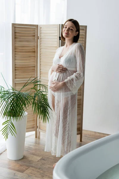 Pleased pregnant woman in robe standing near plant in bathroom