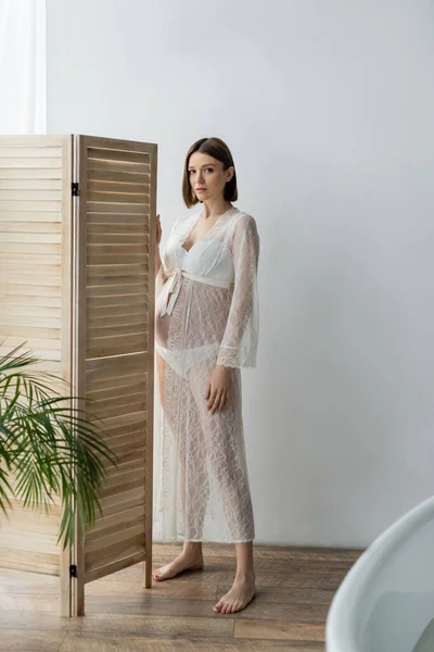 Barefoot pregnant woman in robe standing near folding screen in bathroom
