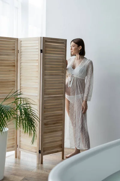 Pregnant woman in robe standing near folding screen and bathtub at home
