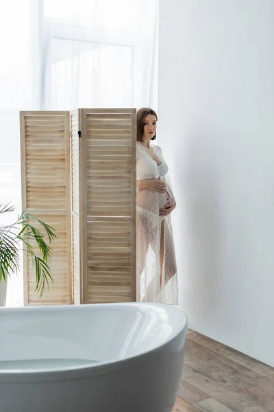 Pregnant woman in robe looking at camera near folding screen and bathtub with water