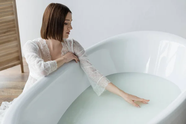 Brunette woman in robe touching water in bathtub at home