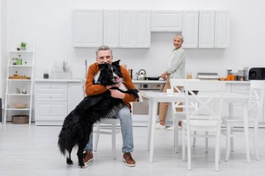 Olgun bir adam mutfakta bulanık karısının yanında sınır köpeğini okşuyor. 