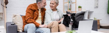 Gülümseyen çift evde dizüstü bilgisayarın yanında çoban köpeğini okşuyor. 