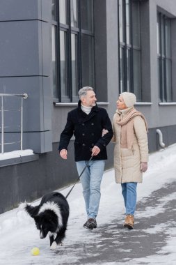 Kış kıyafetli bir çift, şehir sokağında tasma sınırında yürürken konuşuyor. 