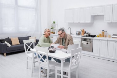 Senior woman talking to husband holding paper near coffee and laptop in kitchen  clipart