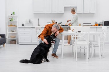 Orta yaşlı bir adam mutfakta kahve fincanının yanında çoban köpeği besliyor. 