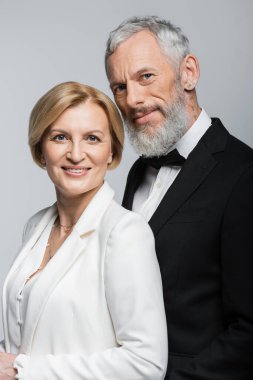 Gülümseyen olgun gelinin portresi. Gri üzerine izole edilmiş takım elbiseli damadın yanındaki kameraya bakıyor. 