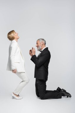 Gri arka planda heyecanlı, olgun bir kız arkadaşın yanında mücevher kutusunu tutan adamın yan görüntüsü 