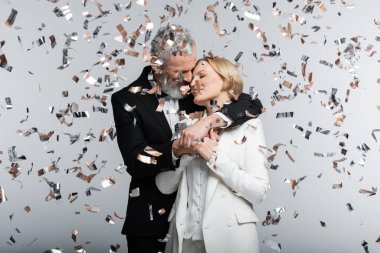 Gri arka planda konfeti altında gelini öpen ve kucaklayan olgun bir damat. 