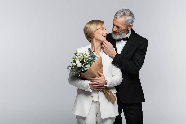 Mature groom touching chin of smiling bride with bouquet isolated on grey 