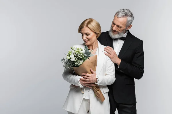 Positive groom in suit hugging middle aged bride with bouquet isolated on grey