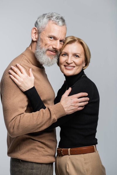 Portrait of trendy mature couple hugging and looking at camera isolated on grey 