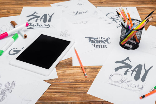 цифровая табличка с чистым экраном, цветные карандаши и бумаги с различными шрифтами на деревянном столе