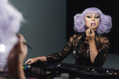drag queen in violet wig and black lace dress applying makeup near mirror clipart