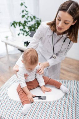 Gülümseyen doktor klinikte çocuğu elektronik ölçekte tutuyor 