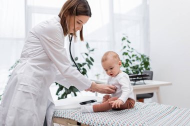 Klinikteki tıbbi kanepede steteskopla bebeği muayene eden genç doktor.  