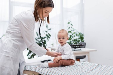 Bebek, klinikte steteskopla gülümseyen doktorun yanındaki kameraya bakıyor. 