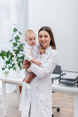 Beyaz önlüklü pozitif aile doktoru kucağında bebekle klinikteki kameraya bakıyor. 