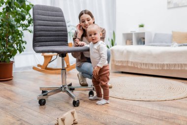 Bebek çocuk koltuğun yanındaki kameraya bakıyor ve yatak odasında gülümseyen anne. 