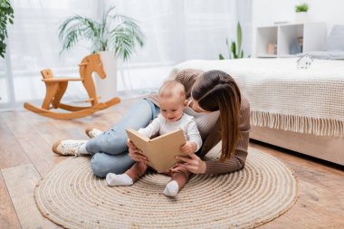 Esmer anne evde halıda neşeli bir bebeğin yanında kitap okuyor. 