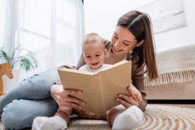 Evde, bebeğin yanında, halıda kitap okuyan, gülümseyen kadın. 