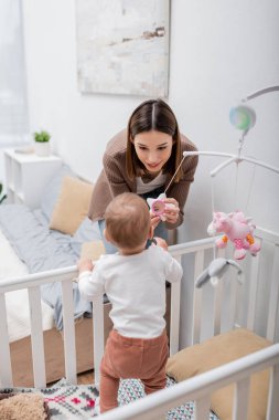 Esmer anne, oğlunun yanında oyuncak tutuyor. Yatak odasında. 