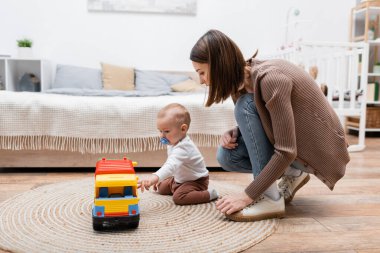 Anne ve babası, evde oyuncak arabasıyla oynayan, emzik takan oğluna bakıyor. 